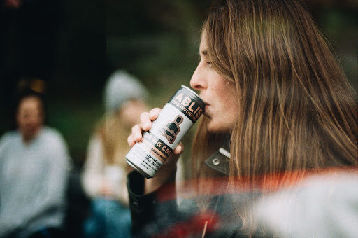 A woman in the foreground drinks a can of Ablis; in the background you can see her friends laughing and talking to each other on Earth Day 2022